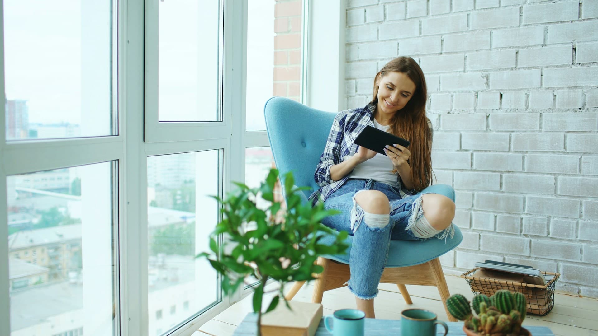 Jovem sorrindo na poltrona da sala do apartamento, com o tablet nas mãos