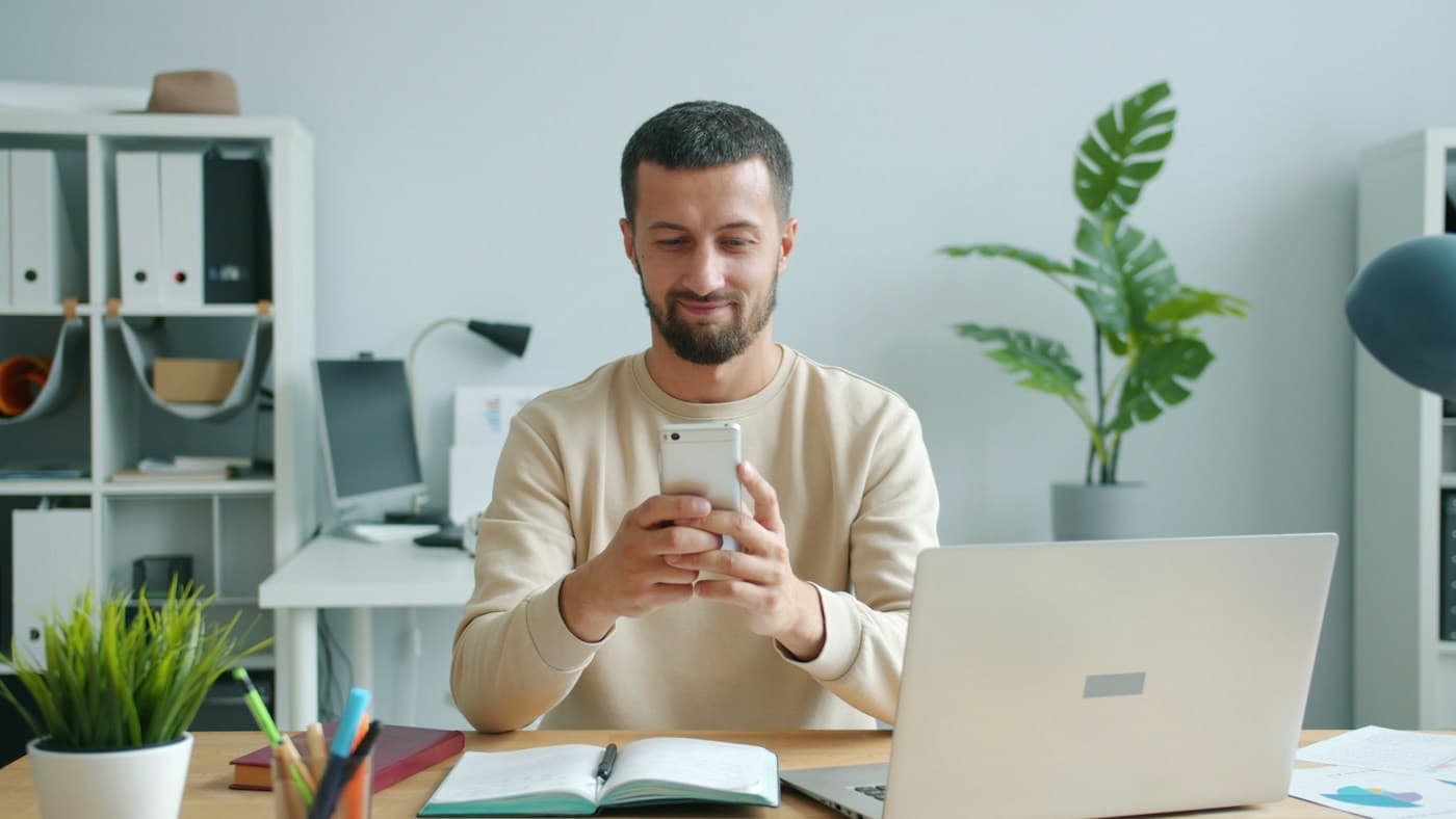 Colaborador sorrindo enquanto usa o celular na sua mesa de trabalho