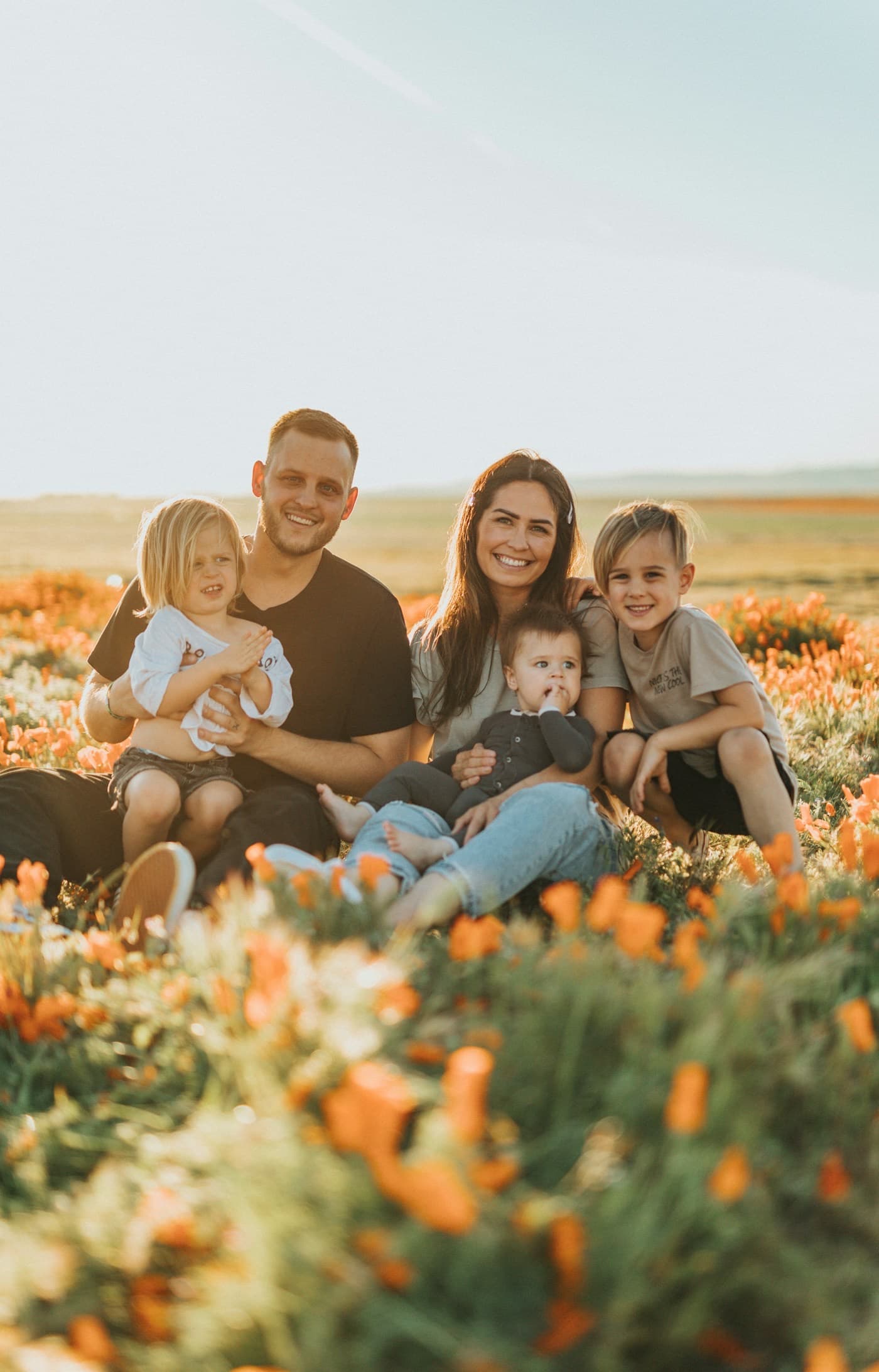Pais sorrindo sentados em um campo florido com os três filhos pequenos